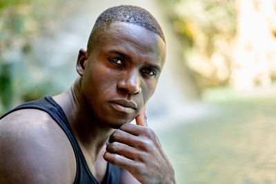 Full body thoughtful black male in casual clothes touching face and looking at camera while sitting on rock near clean lake on summer day