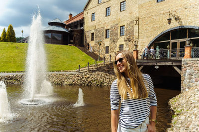 A long-haired woman in a striped jumper and sunglasses laughs next to a fountain in the summer