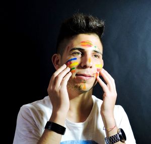 Close-up of young man applying colorful paint on face against black wall