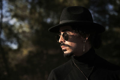 Side view of young hipster bearded male traveler with piercing wearing stylish black hat and sunglasses looking away while standing on blurred countryside