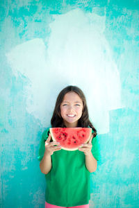 Portrait of happy girl having watermelon while standing by turquoise wall