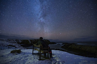 Rear view of man sitting on bench against sky at night