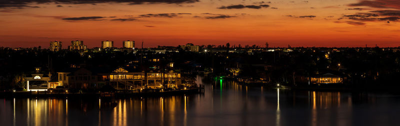 Illuminated buildings by river against sky at sunset