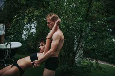 Side view of shirtless young man and plants against trees