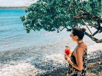 Young woman drinking water at sea shore against sky