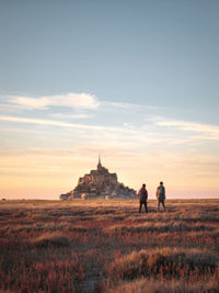 Rear view of hikers looking at view of mont saint-michel against cloudy sky during sunset