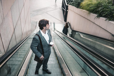 High angle view of businessman holding coffee standing on escalator smiling against sky