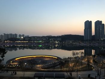 Illuminated city by river against sky during sunset