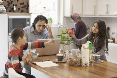 Muslim man working on laptop in kitchen with family having breakfast