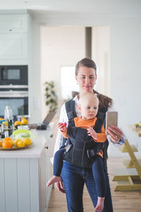 Female freelancer blogging while carrying daughter in kitchen using smart phone at home