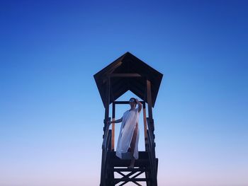 Low angle view of woman standing on lifeguard hut against clear blue sky during sunset