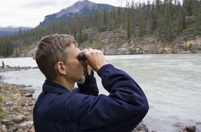 Mature man looking through binoculars while standing at lakeshore