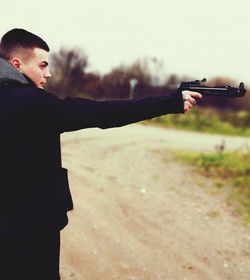 Side view of man aiming gun while standing on dirt road