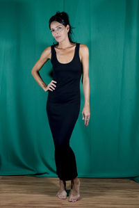 Portrait of standing adult model in black dress against dark green background. 