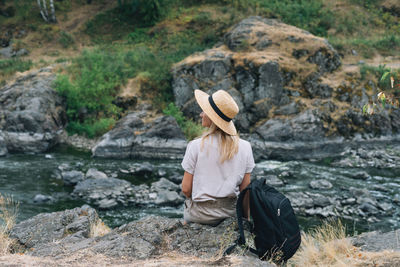 Young woman traveler in straw hat with backpack sits and looks at the mountain river, local travel 