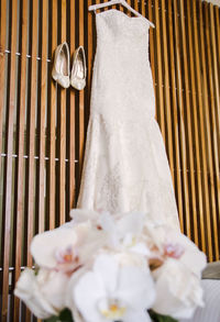 Wedding dress hanging on wood next to shoes with bouquet flowers out of focus in the foreground