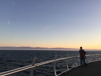 Man standing on railing by sea against clear sky