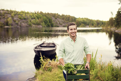 Smiling man carrying vegetables crate while standing against boat at lake