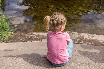 A little girl sits with her back to a pond in the park.