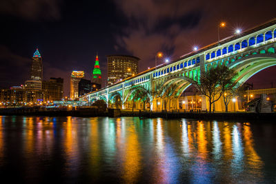 Illuminated bridge over river against buildings at night