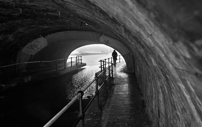 Rear view of silhouette person walking on footpath by river in tunnel