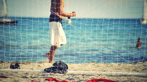 Low section of shirtless man standing at beach seen through net