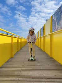 Little girl with a helmet on a kick scooter yellow walls blue sky