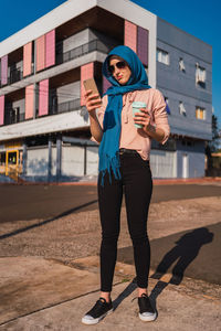 Trendy arab female in traditional hijab standing with coffee to go in street and messaging on cellphone on sunny day in city