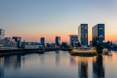 Illuminated buildings by river against sky during sunset