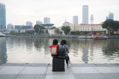 Rear view of people sitting on riverbank against cityscape