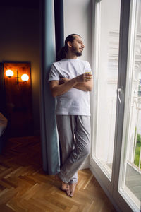 Young man with a beard in a t-shirt and trousers barefoot stands with orange juice at the window 