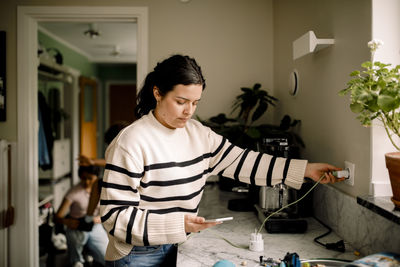 Woman charging smart phone while standing near kitchen counter at home