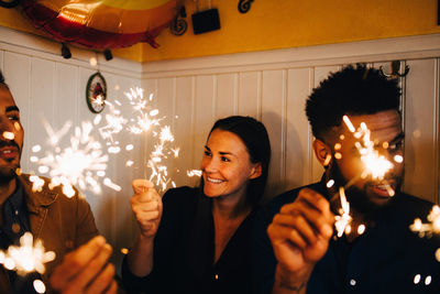 Smiling young woman with multi-ethnic male friends holding sparklers in restaurant during dinner party