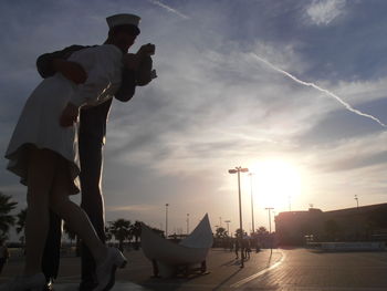People on sidewalk in city against sky during sunset