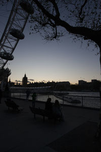 Silhouette people sitting on riverbank against sky during sunset