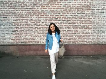 Portrait of young woman walking on footpath against brick wall