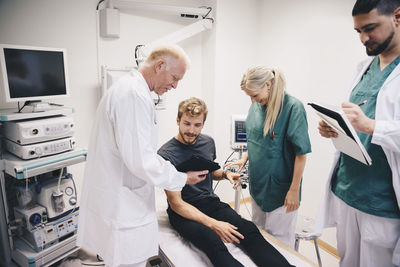 Mature doctor showing digital tablet to young male patient on bed by colleagues in hospital