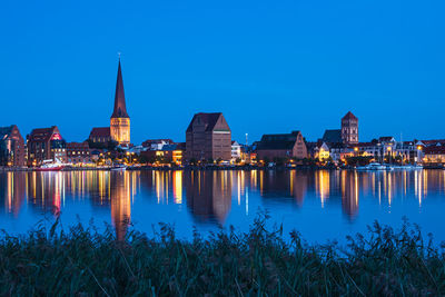 Illuminated buildings by river against blue sky at night