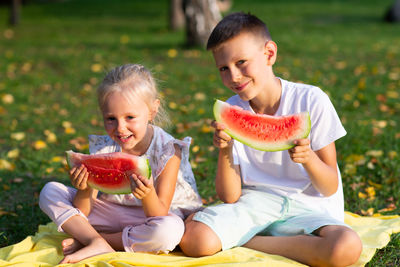 Brother and sister holding watermelon while sitting in park