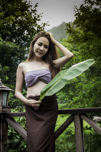 Beautiful young woman standing by railing against trees