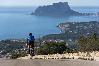 Rear view of man riding bicycle on road by sea