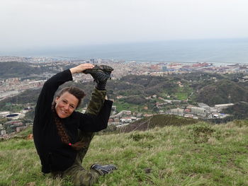 Portrait of smiling woman stretching on grass at mountain peak against clear sky