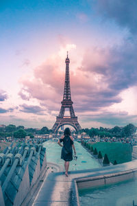 Rear view of woman looking at eiffel tower while standing on wall against cloudy sky