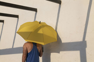 Rear view of adult man holding yellow umbrella against white wall with shadow in summer