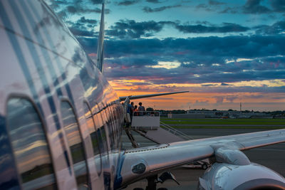 Passengers on boarding bridge at airport runway against sky during sunset