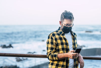 Mature man wearing mask using smart phone while standing against sea