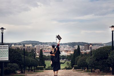 Rear view of young woman in graduation gown holding slate while standing on footpath against cloudy sky
