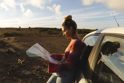 Woman with map leaning on car at sunset