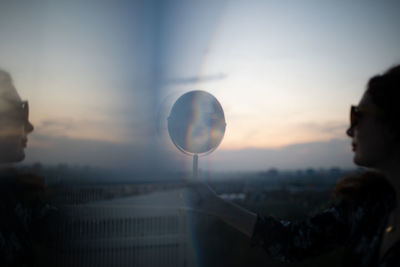 Side view of woman holding mirror while standing against sky during sunset