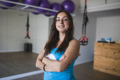 Positive young female yoga trainer in activewear standing with arms crossed and looking at camera against blurred interior of fitness studio